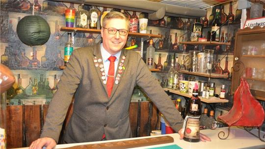 Mann in grauem Sakko mit roter Krawatte und Anstecknadeln steht hinter einer Bar mit Flaschen und Gläsern im Hintergrund