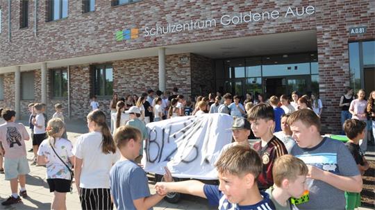 Gruppe von Kindern und Jugendlichen vor einem Backsteingebäude mit der Aufschrift 'Schulzentrum Goldene Aue'