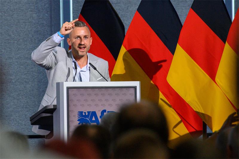 Ein Mann steht an einem Rednerpult und spricht, im Hintergrund Deutschlandflaggen.