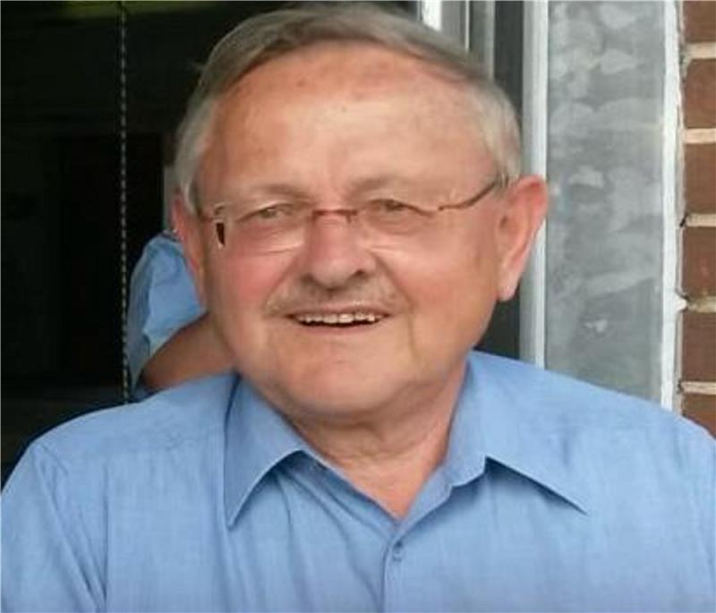 Älterer Mann mit grauen Haaren trägt ein hellblaues Hemd und steht vor einer Wand mit einem Fenster und Ziegelsteinen.