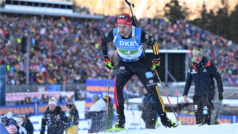 Biathlet mit Startnummer 4 in schwarzer Sportkleidung und blauem Trikot beim Wettkampf auf Schnee vor großem Publikum im Hintergrund