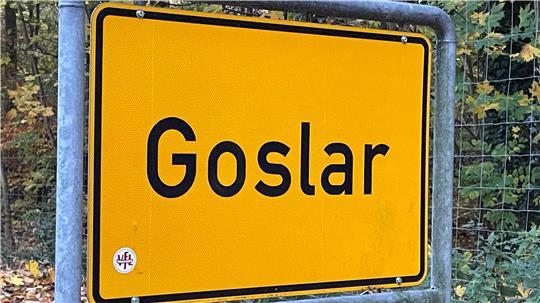 Gelbes Ortsschild mit schwarzer Umrandung und der schwarzen Aufschrift 'Goslar' vor einem Waldzaun