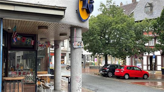 Straßenszene mit überdachtem Gehweg, mehreren Autos, Bäumen und Gebäuden im Hintergrund an einem regnerischen Tag
