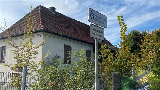 Haus mit rotem Ziegeldach hinter grünem Buschwerk und zwei Verkehrsschildern mit Texten