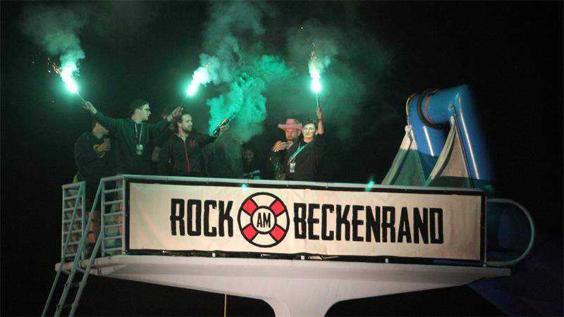 Menschen halten grüne Leuchtfackeln in die Luft, während sie auf dem Sprungturm des Freibads stehen. Auf einem Banner steht "Rock am Beckenrand".