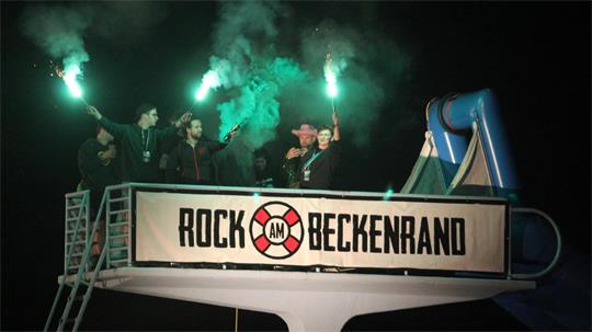 Menschen halten grüne Leuchtfackeln in die Luft, während sie auf dem Sprungturm des Freibads stehen. Auf einem Banner steht "Rock am Beckenrand".