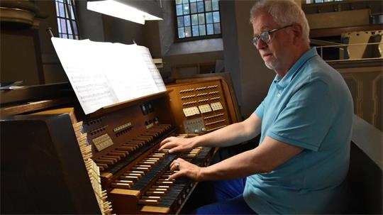 Älterer Mann mit Brille spielt an einer großen Orgel in einem Raum mit hohen Fenstern.