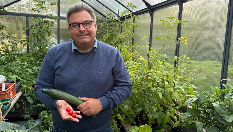 Ein Mann steht in einem Gewächshaus und hält Tomaten und eine Gurke in der Hand. 