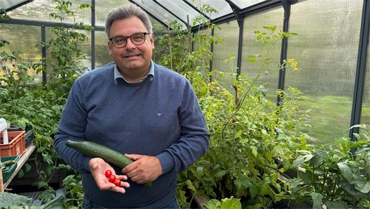 Ein Mann steht in einem Gewächshaus und hält Tomaten und eine Gurke in der Hand. 