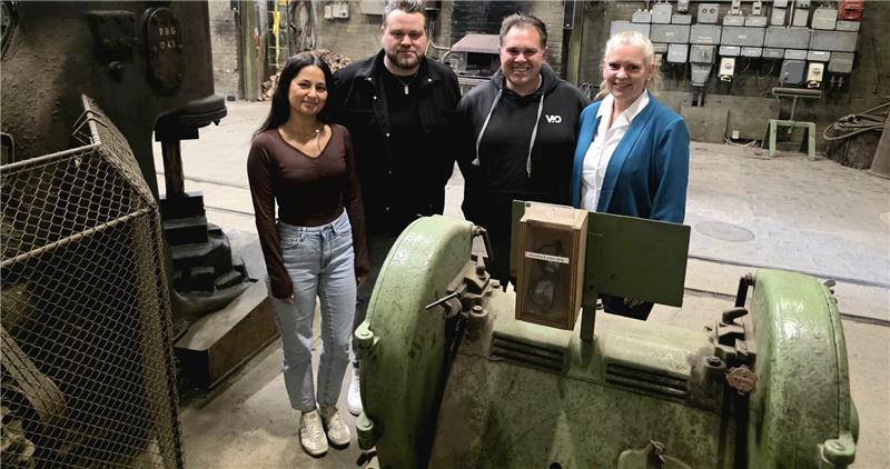 Vier Personen stehen in einer alten Industriehalle neben großen grünen Maschinen und elektrischen Schaltkästen an der Wand