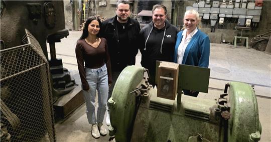 Vier Personen stehen in einer alten Industriehalle neben großen grünen Maschinen und elektrischen Schaltkästen an der Wand