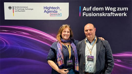 Zwei Personen mit Namensschildern vor einem violetten Hintergrund mit den Logos des Bundesministeriums für Forschung, Technologie und Raumfahrt sowie der Hightech Agenda Deutschland und dem Text 'Auf dem Weg zum Fusionskraftwerk
