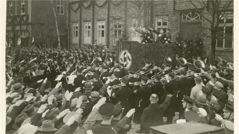 Große Menschenmenge vor einem Gebäude, die mit ausgestrecktem Arm einen Hitlergruß zeigt, während mehrere Musiker in Uniform auf der rechten Seite spielen