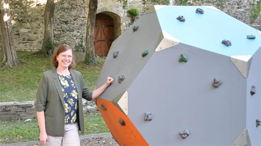 Frau steht lächelnd neben einem großen, mehrfarbigen, polygonalen Kletterwürfel im Freien vor einer Steinmauer