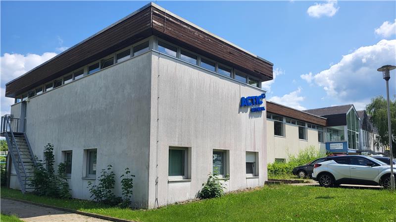 Zweistöckiges Gebäude mit weißer Fassade, braunem Dach und blauer Schrift auf der Vorderseite, davor Rasen und geparkte Autos