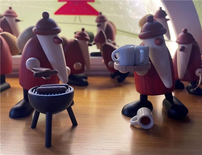 Heiße Geschenktipps: Kleine weißbärtige Rotnasen aus Sachsen Mehrere hölzerne Weihnachtsmänner mit roten Mänteln und weißen Bärten, einer hält Tassen auf einem Tablett in der Hand, ein anderer steht neben einem kleinen Grill, auf einem Holztisch.