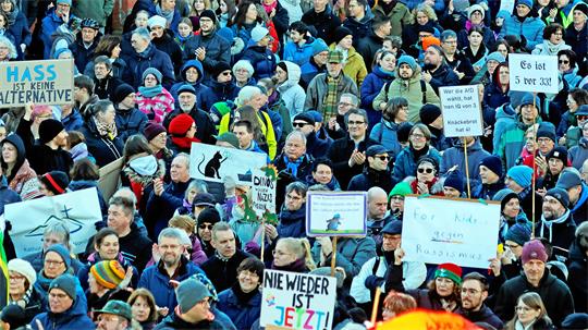 Menschenmenge bei einer Demonstration mit verschiedenen Schildern und Plakaten, darunter eines mit der Aufschrift 'NIE WIEDER IST JETZT'