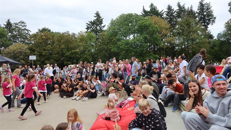 Große Menschenmenge sitzt und steht im Freien vor einer Gruppe von Kindern, die sich bewegen, umgeben von Bäumen