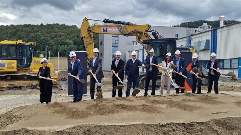 Gruppe von neun Personen mit Schutzhelmen steht mit Schaufeln auf einer Baustelle vor einem Bagger und hebt gemeinsam Sand auf.