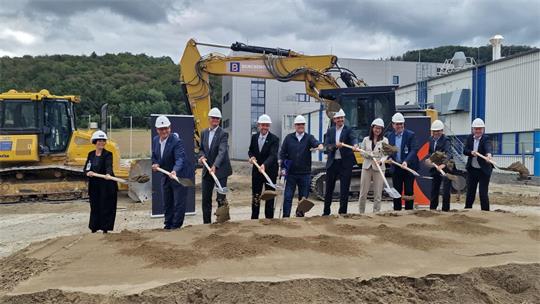 Gruppe von neun Personen mit Schutzhelmen steht mit Schaufeln auf einer Baustelle vor einem Bagger und hebt gemeinsam Sand auf.