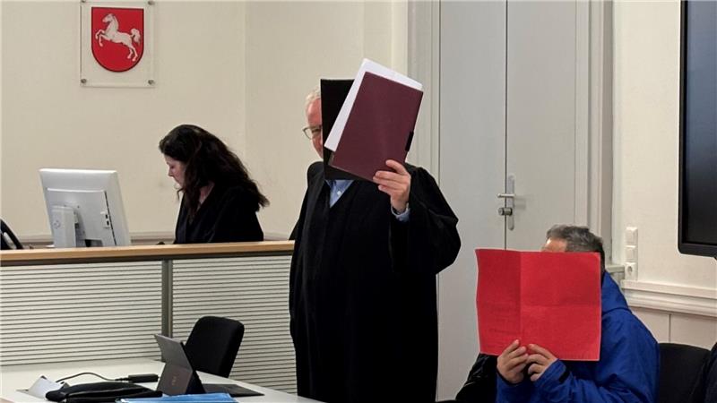 Person in Anwaltsrobe hält ein Buch hoch, daneben sitzt eine Person mit blauem Pullover, die ein rotes Buch vor das Gesicht hält, im Hintergrund eine Person an einem Computer.