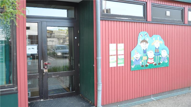 Eingangstür eines Gebäudes mit Glasfenstern, daneben bunte Wandmalerei mit fünf kindlichen Figuren und zwei Informationsplakaten.