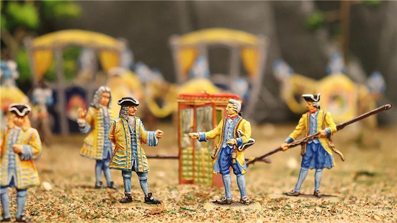 Perücken, Pomp und Pulverdampf im Goslarer Zinnfigurenmuseum Mehrere Miniaturfiguren in historischer Kleidung umgeben eine kleine, gelbe Sänfte auf einem sandigen Untergrund