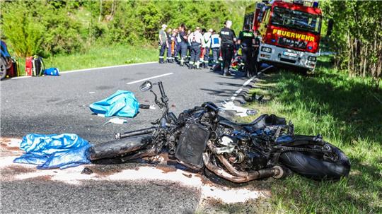 Ein zerstörtes Motorrad nach einem Unfall auf einer Straße, im Hintergrund sind Rettungskräfte und ein Feuerwehrfahrzeug zu sehen.