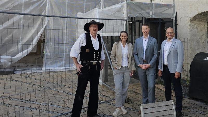 Ein Zimmerer in alter Kluft steht neben einer Frau und zwei Männern in Jackets vor einer Baustelle.