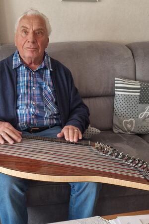 Älterer Mann sitzt auf einem Sofa und hält ein Zitherinstrument auf dem Schoß.