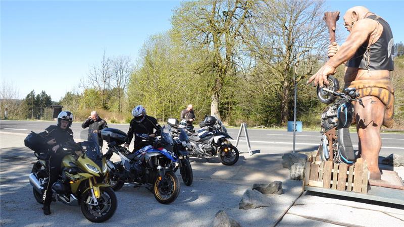 Mehrere Motorradfahrer mit Helmen auf parkenden Motorrädern auf einem Parkplatz neben einer großen Statue eines nackten Mannes mit Keule