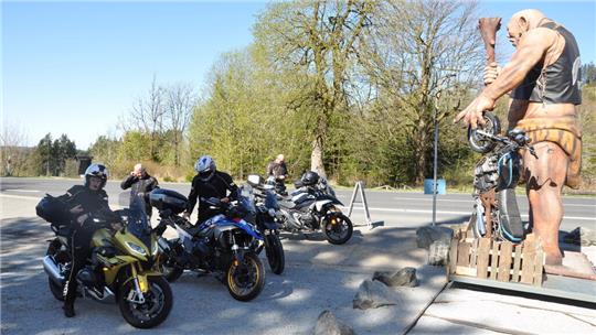 Mehrere Motorradfahrer mit Helmen auf parkenden Motorrädern auf einem Parkplatz neben einer großen Statue eines nackten Mannes mit Keule