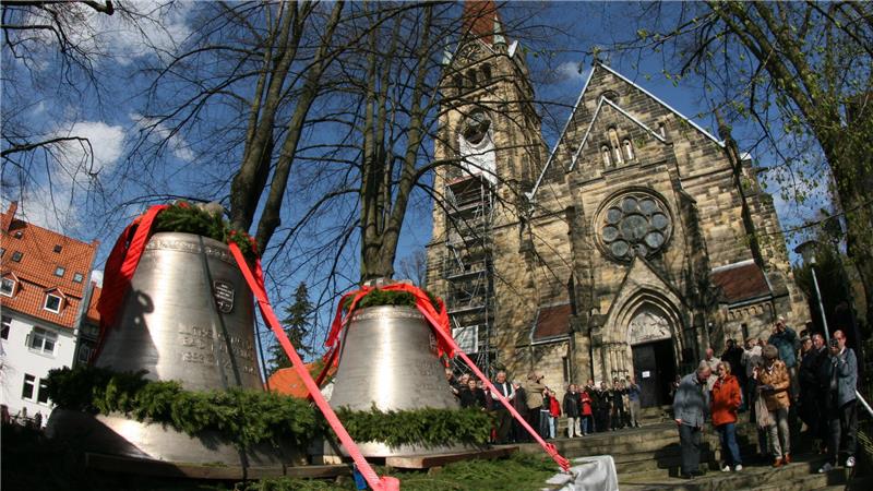 Zwei Glocken stehen vor einer Kirche, während Menschen drumherum stehen.