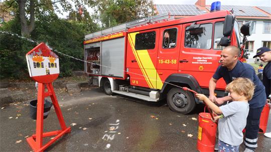 Ein Junge und ein Mann an einer Kübelspritze, im Hintergrund ein Feuerwehrauto.