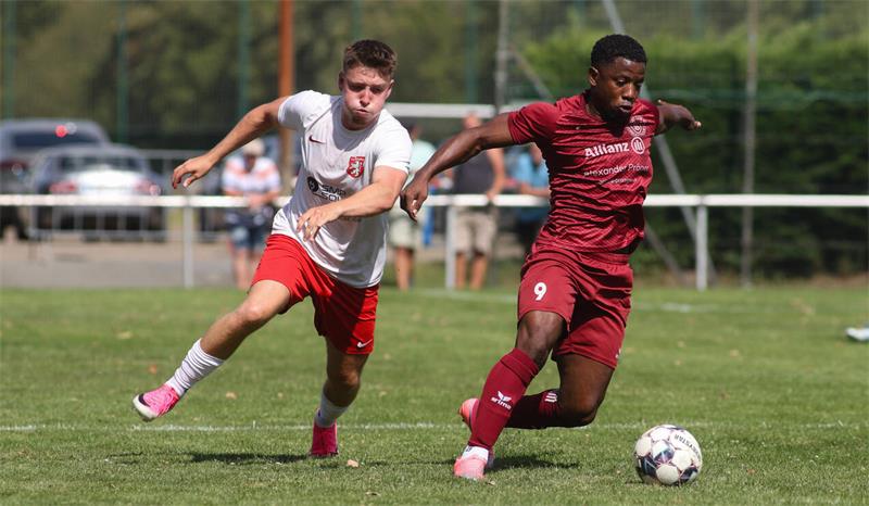 Zwei Fußballspieler in Aktion auf dem Spielfeld, einer in weißem Trikot und roter Hose, der andere in rotem Trikot und Hose, beide laufen dem Ball hinterher