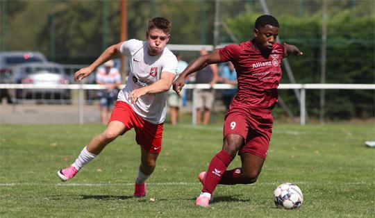 Zwei Fußballspieler in Aktion auf dem Spielfeld, einer in weißem Trikot und roter Hose, der andere in rotem Trikot und Hose, beide laufen dem Ball hinterher