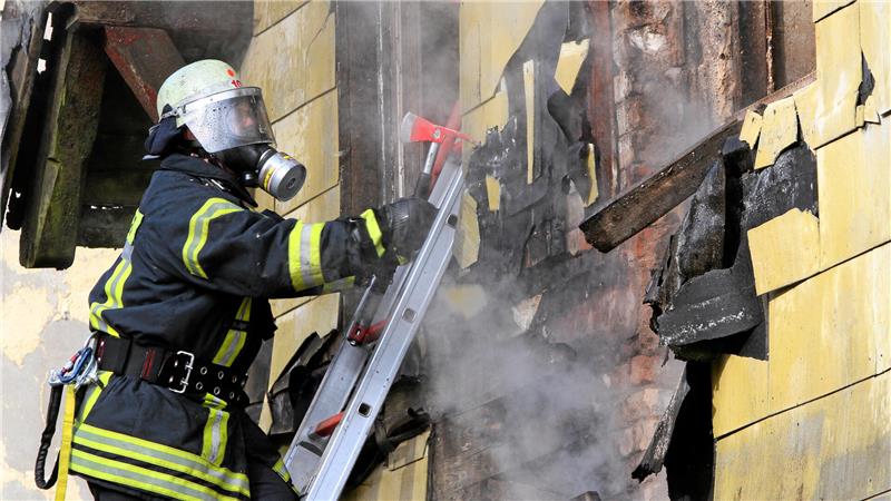 Feuerwehrmann in Schutzkleidung und Atemschutzgerät auf Leiter löscht brennende, beschädigte Hauswand mit Axt in der Hand