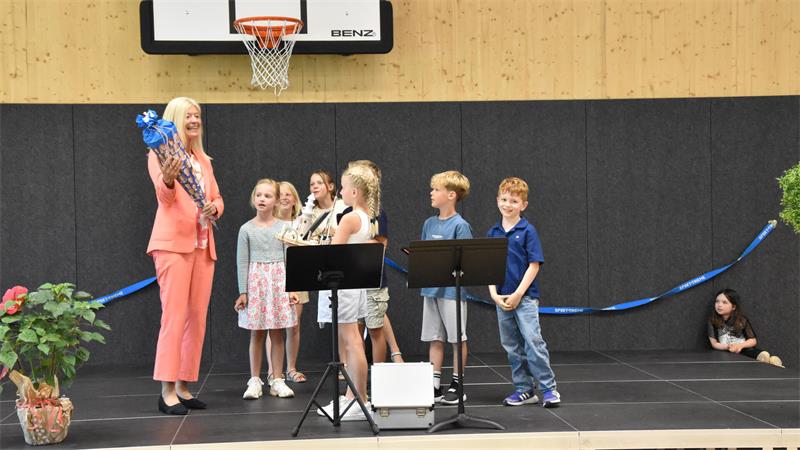 Frau in korallfarbenem Anzug hält Schultüte vor Gruppe von Kindern auf Bühne in Turnhalle mit Basketballkorb.