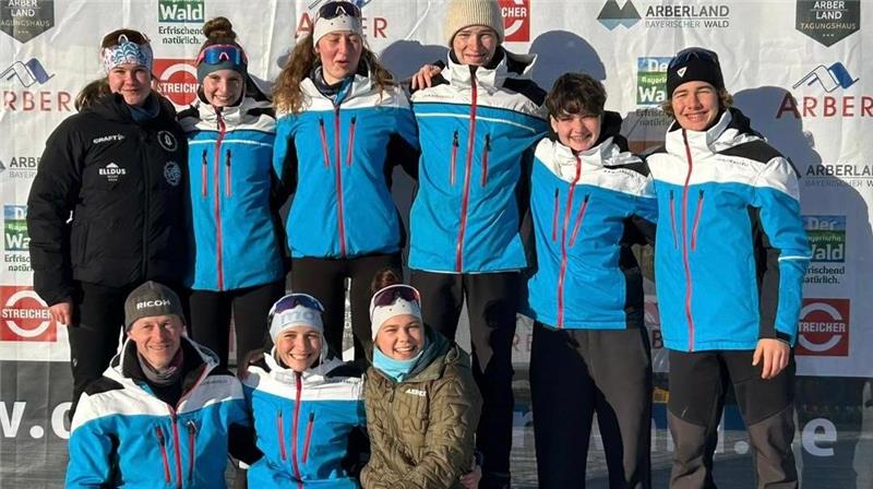 Gruppe von neun Personen in blauen und schwarzen Sportjacken steht und sitzt auf einem Podest mit den Zahlen 1 und 3, im Hintergrund Werbetafeln mit Logos und Texten.