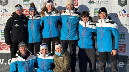 Gruppe von neun Personen in blauen und schwarzen Sportjacken steht und sitzt auf einem Podest mit den Zahlen 1 und 3, im Hintergrund Werbetafeln mit Logos und Texten.