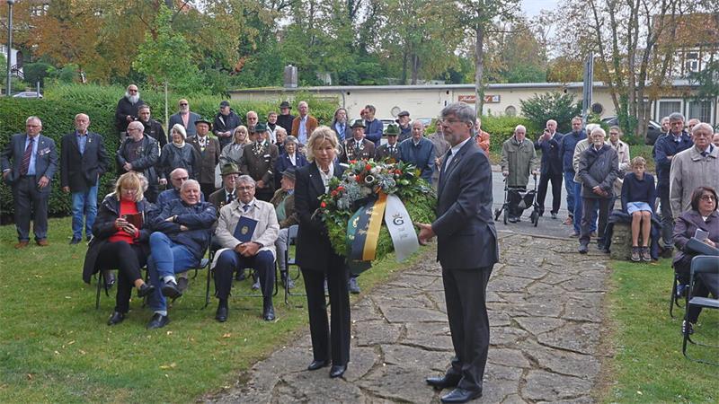 Zwei Personen tragen einen Kranz vor einer sitzenden und stehenden Menschenmenge im Freien