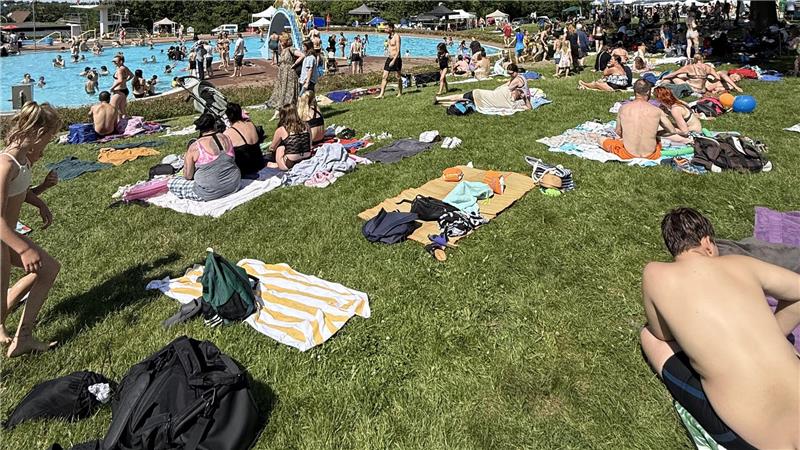 Menschen liegen und sitzen auf Handtüchern auf einer Wiese neben einem Schwimmbecken bei sonnigem Wetter
