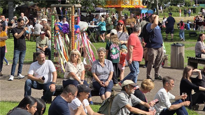 Menschen sitzen und stehen auf einer Wiese und einem gepflasterten Weg bei einem Fest mit buntem Karussell und Ständen im Hintergrund.