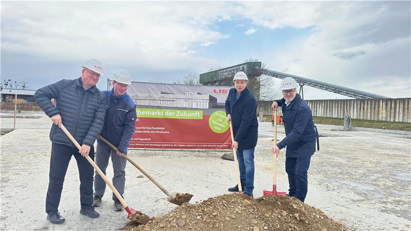 Vier Männer mit Schutzhelmen und Winterjacken schaufeln gemeinsam Erde auf einer Baustelle vor einem großen Schild