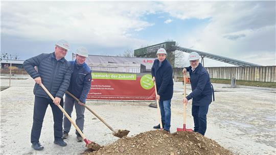 Vier Männer mit Schutzhelmen und Winterjacken schaufeln gemeinsam Erde auf einer Baustelle vor einem großen Schild