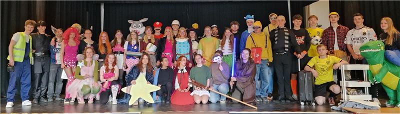 Große Gruppe von Menschen in bunten Kostümen auf einer Bühne, einige halten Requisiten wie einen Stern und eine Leiter.