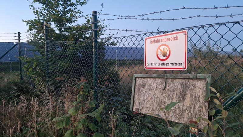 Drahtzaun mit Stacheldraht und einem Warnschild mit der Aufschrift 'Gefahrenbereich Zutritt für Unbefugte verboten' vor einer bewachsenen Fläche und blauem Himmel.