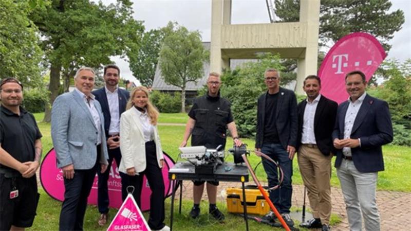 Gruppe von acht Personen steht im Freien vor einem Tisch mit technischer Ausrüstung und Telekom-Logos auf Fahnen im Hintergrund.