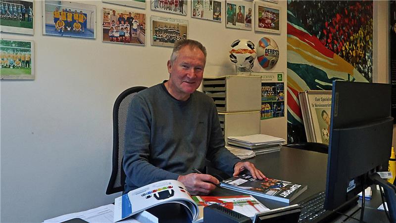 Mann sitzt an Schreibtisch mit geöffneten Büchern, im Hintergrund viele gerahmte Fotos und ein Fußball auf einem Schrank.