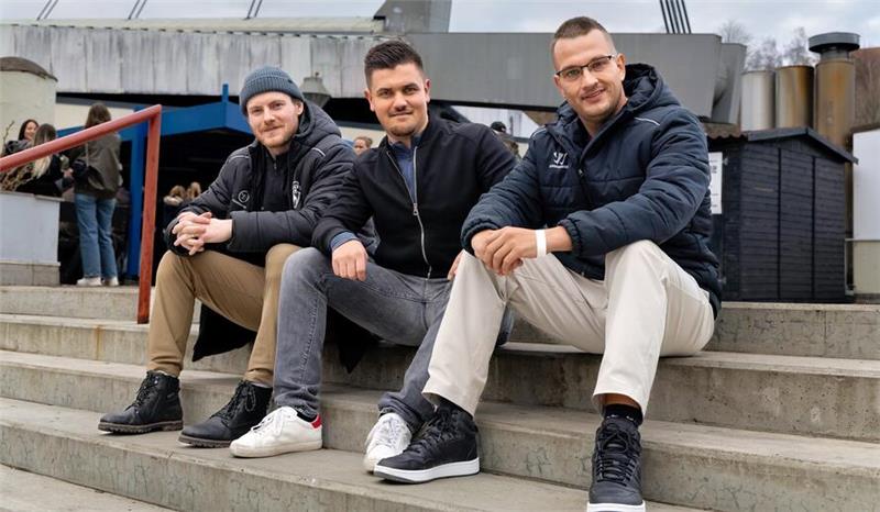 Drei Männer sitzen auf einer Treppe vor einer Brücke und tragen Freizeitkleidung mit Jacken und Sneakers